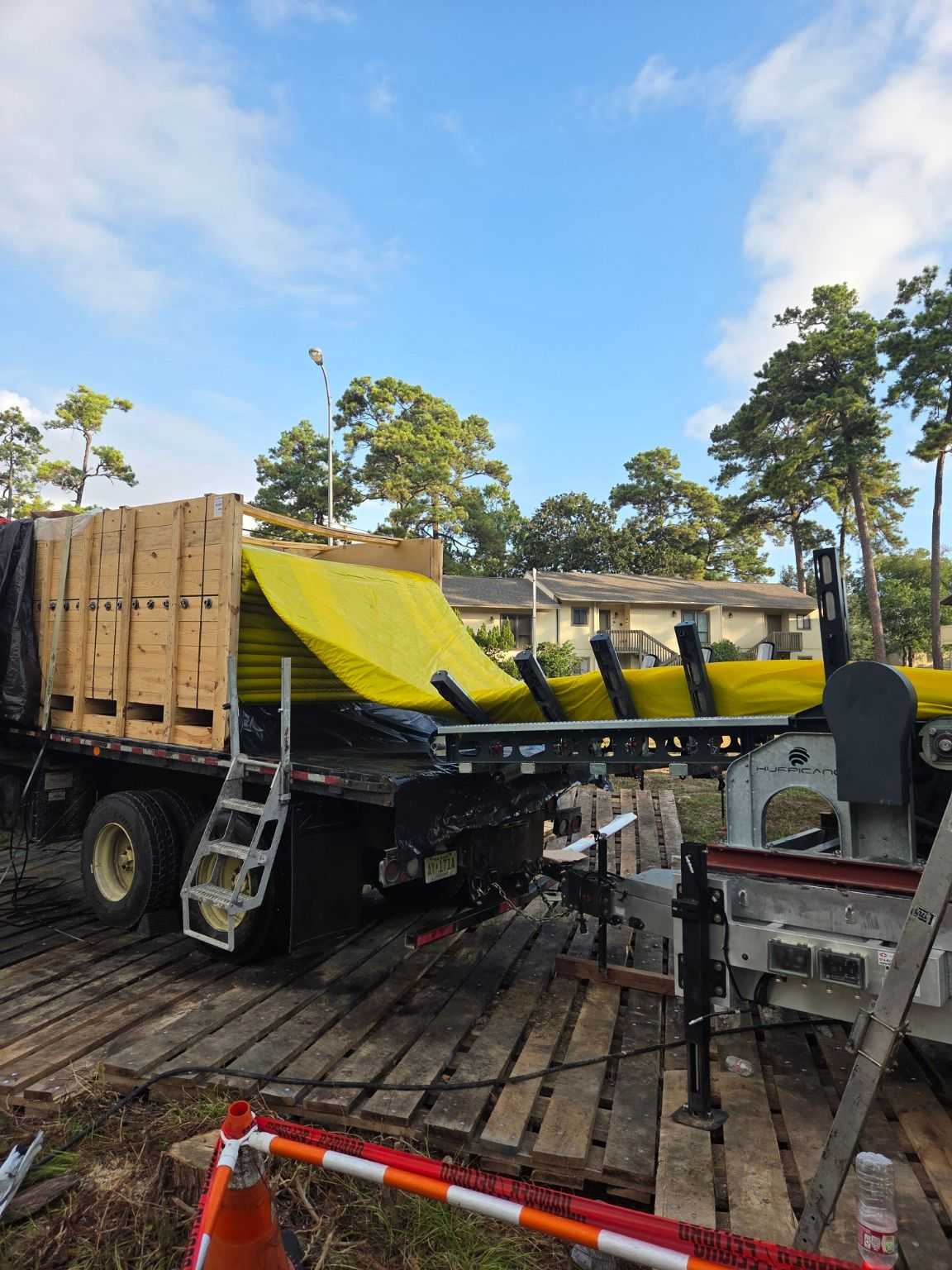 Utility construction equipment unloading large yellow pipe sections from a flatbed truck at a work site, with traffic barriers, ladders, and nearby residential buildings in the background.