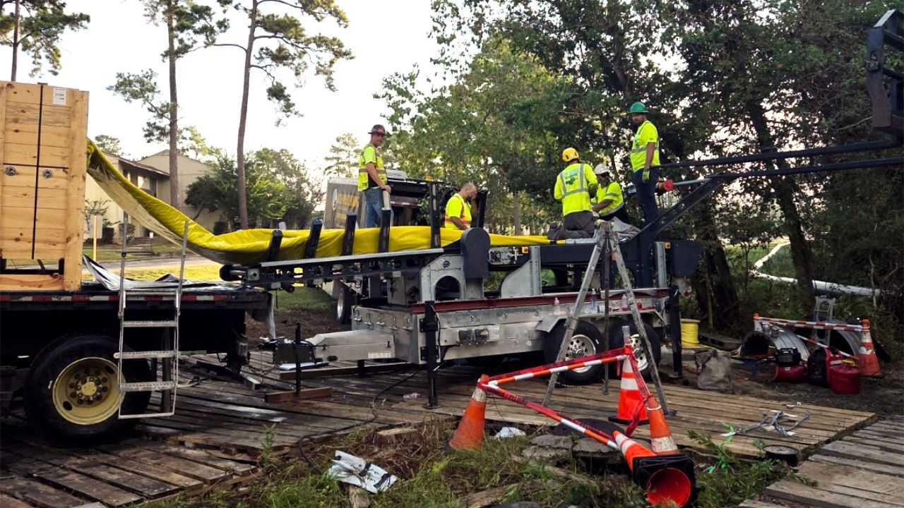 Construction crew installing utility infrastructure at a roadside work site, using specialized equipment, traffic cones, trucks, and safety gear near a wooded area.
